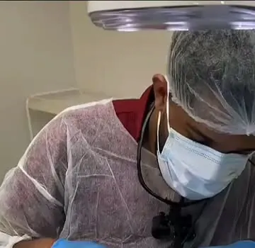 Maylson, de touca, máscara e lupas, atende um paciente na cadeira odontológica