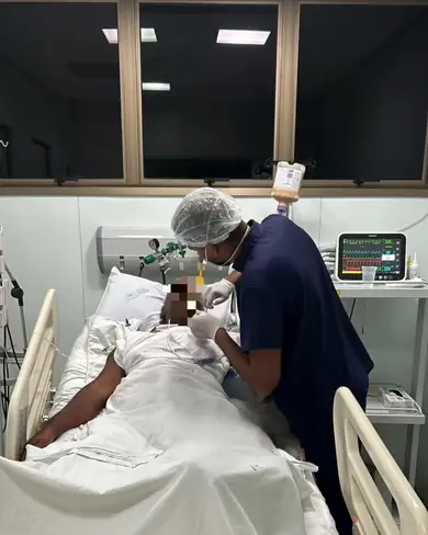 Maylson, de touca e jaleco, atende um paciente internado em um leito de UTI