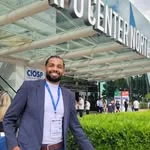 Maylson Cruz sorrindo, de terno e crachá, em frente a um centro de convenções